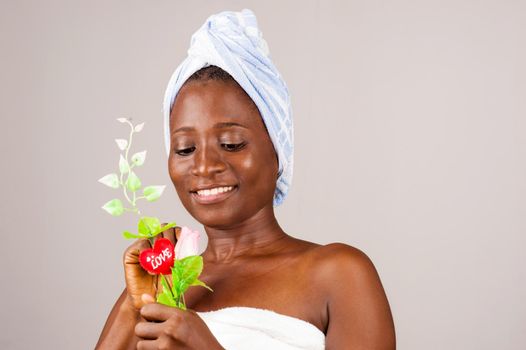 African girl standing on gray background, bath towel knotted on the head while admiring a flower smiling.