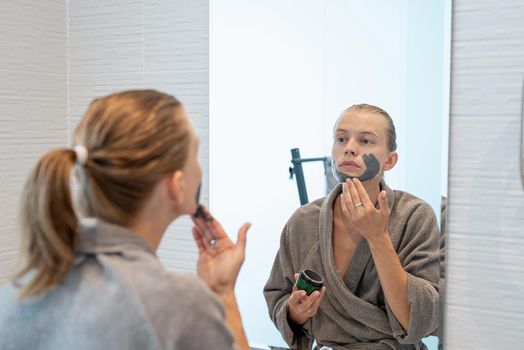 Womens health. Spa and wellness. Woman in gray bathrobes applying face mask in the bathroom looking at the mirror