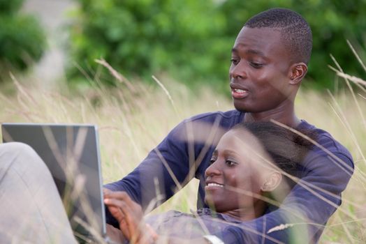 young african couple sitting in the grass with laptop and look at the screen smiling.
