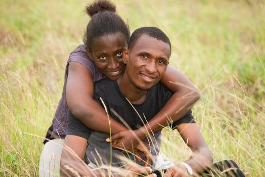 young african couple sitting in an embracing park and looking at camera smiling.