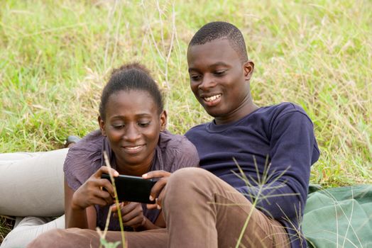 young african couple intertwined in a park looking at mobile phone while smiling.