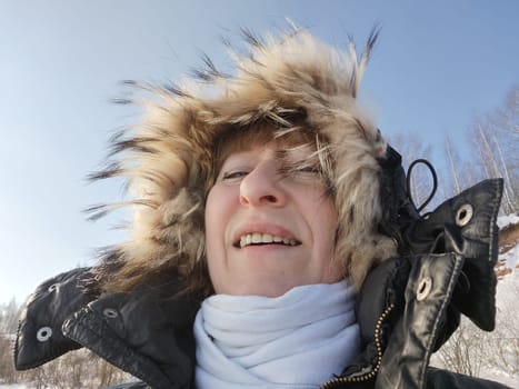 A cheerful girl or woman in a hooded jacket with fur on a sunny spring or winter day. An adult lady on cold day