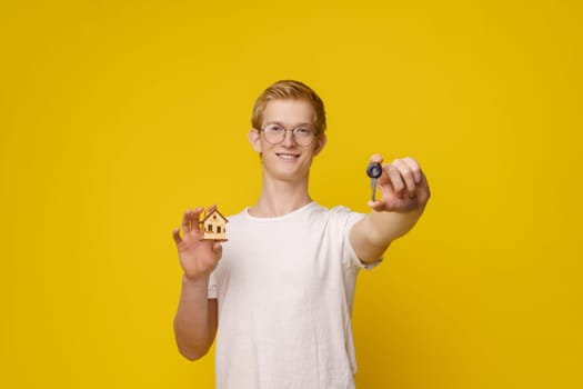 Young man stands against vibrant yellow background, holding set of keys and model house, symbolizing home ownership and the real estate industry. Focus on hand with key. High quality photo