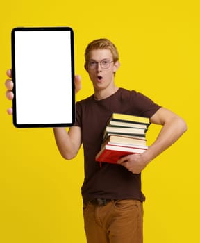 Surprised student holding books and a Tablet PC with a blank white screen for copy space against a yellow background, showcasing the concept of modern education and the integration of technology in learning and studying. High quality photo