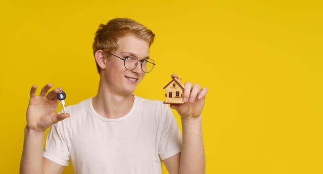 Young man holding a home architectural model, maquette and key against yellow background, showcasing the achievement of home ownership and success in the real estate market. High quality photo
