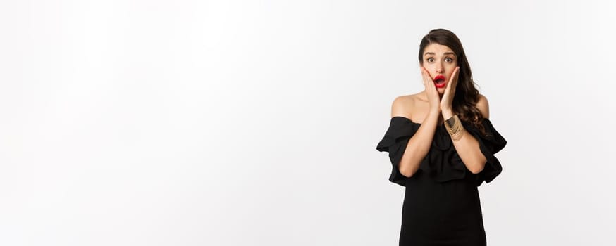 Beauty and fashion concept. Waist-up shot of surprised woman in black cocktail dress, looking amazed at camera, open mouth fascinated, standing over white background.