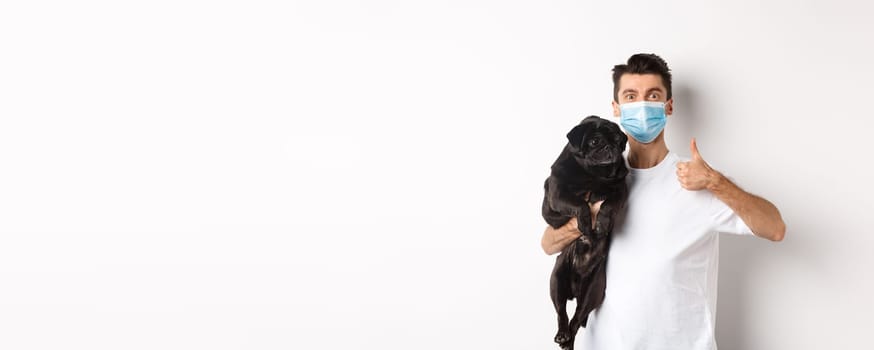 Covid-19, animals and quarantine concept. Young man in medical mask holding cute black pug dog, showing thumb up, like and approve, standing over white background.