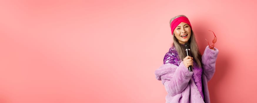 Gorgeous asian senior woman singing karaoke in mic, perform song and looking happy, standing over pink background.