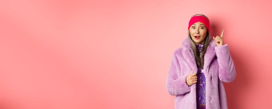 Fashion and shopping concept. Smiling elderly woman having an idea. Stylish old lady in purple fake fur coat raising index finger, suggesting plan, standing excited on pink background.