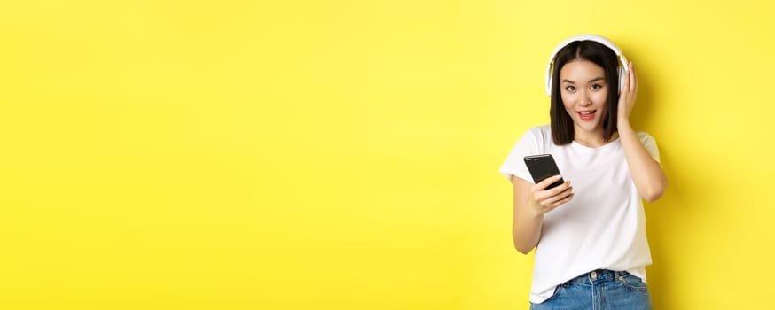 Modern asian woman listening music in wireless headphones, reading smartphone screen and smiling, standing in white t-shirt over yellow background.