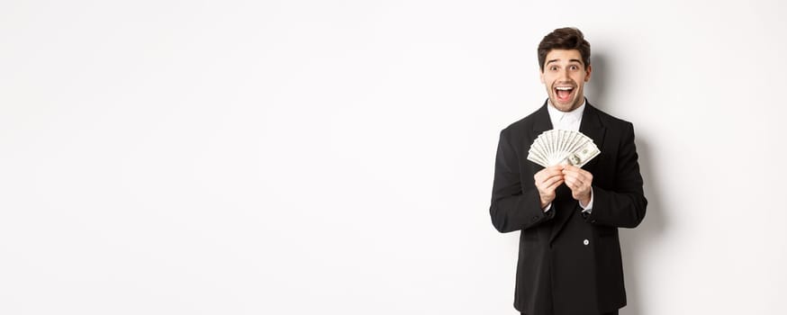 Portrait of handsome successful businessman in trendy suit, showing money and smiling amazed, earn cash, standing over white background.
