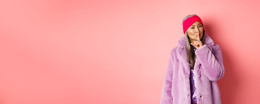 Fashion and shopping concept. Modern chinese senior woman shushing, telling a secret and looking cunning at camera, standing in trendy fake fur coat and headband on pink background.