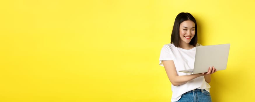 Cute asian female student working on laptop, reading screen and smiling, yellow background.