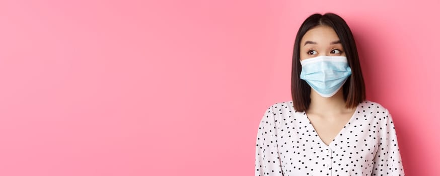 Covid-19, pandemic and lifestyle concept. Beautiful asian female model in medical mask looking left at copy space with surprised face, standing over pink background.