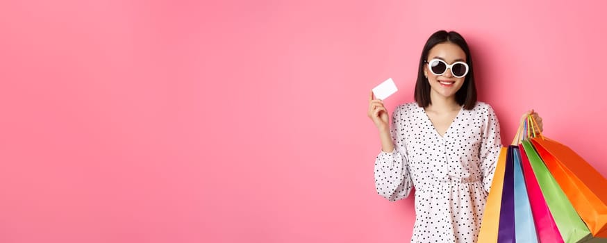 Beautiful asian woman in sunglasses going shopping, holding bags and showing credit card, standing over pink background.