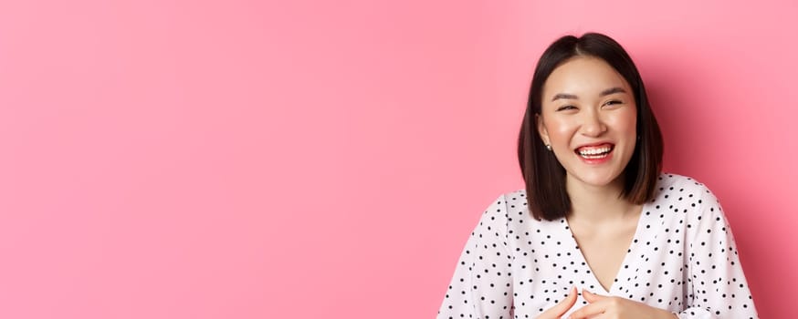 Beauty and lifestyle concept. Close-up of happy asian woman laughing and having fun, standing over pink background. Copy space