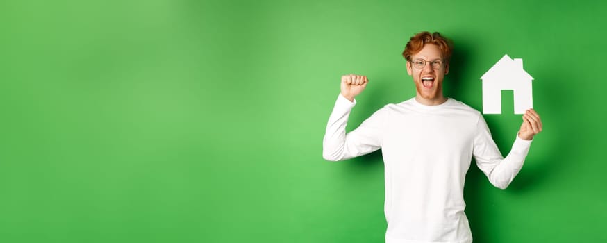 Real estate. Happy caucasian man in glasses with red hair celebrating, showing paper house cutout and making fist pump sign, shouting for joy, buying property, green background.