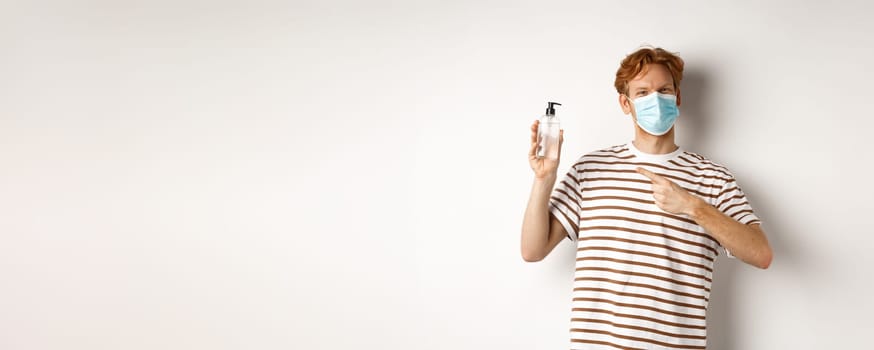 Covid-19, health and lifestyle concept. Cheerful redhead man in face mask pointing finger at hand sanitizer, recommending antiseptic, white background.