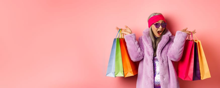 Stylish asian senior woman going shopping, wearing trendy clothes and sunglasses, holding store bags with gifts, treat yourself concept, pink background.