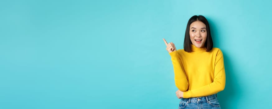 Shopping concept. Portrait of attractive korean girl in yellow sweater, showing promotion offer on copy space, pointing and looking left with pleased smile, blue background.
