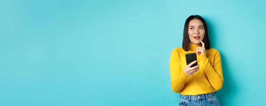 Pensive asian girl holding smartphone and thinking what to order online, standing over blue background.