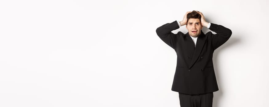 Frustrated and worried businessman in black suit, panicking as looking at trouble, holding hands on head alarmed, standing against white background.