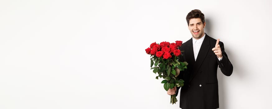Image of handsome romantic guy in black suit, holding bouquet of roses and pointing at camera, congratulating with holiday, standing against white background.