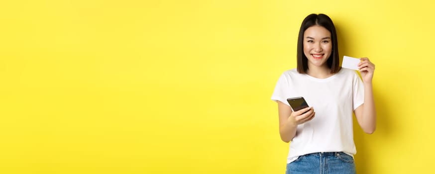 Attractive korean woman paying online with smartphone, showing plastic credit card and smiling, standing over yellow background.