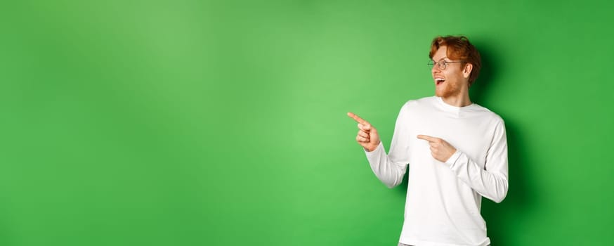 Surprised redhead man in glasses checking out promo, pointing and looking left with amazed face, standing in long-sleeve t-shirt over green background.