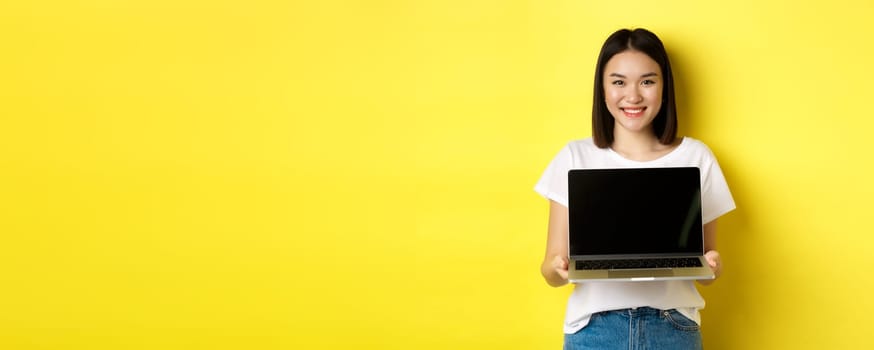 Young asian woman demonstrate online offer, showing laptop screen and smiling, standing over yellow background.