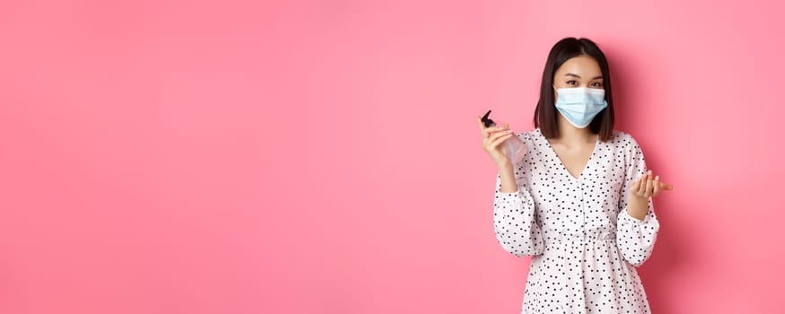 Covid-19, pandemic and lifestyle concept. Cute asian woman clean hands with sanitizer, using antiseptic and wearing face mask, standing over pink background.