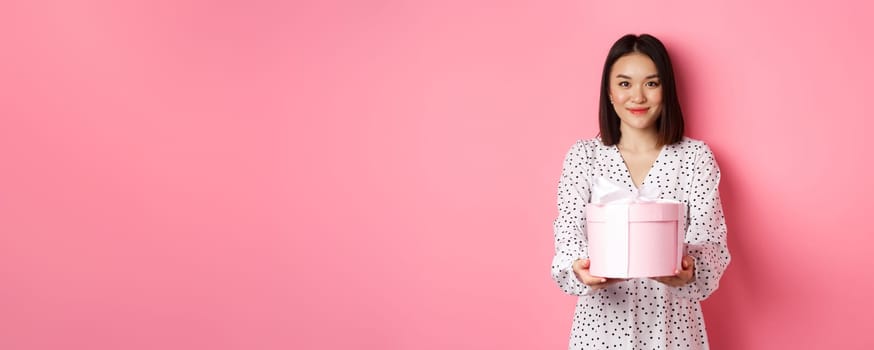 Beautiful asian woman wishing happy holidays, giving you gift in cute box, standing against pink background.