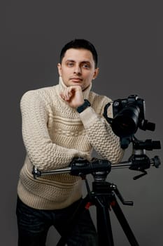 Professional young handsome photographer in sweater with dslr photo camera on tripod on gray background