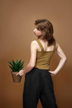 young woman with stylish hairstyle posing with plant in pot on beige background.