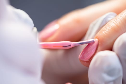 Maintaining the principles of hygiene in the beauty salon. The beautician holds the client's index finger in her hands. A close-up of the nails being painted. Performing a hybrid manicure service.