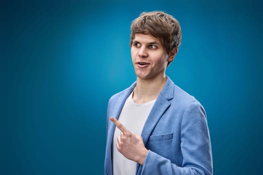 caucasian young man in jacket pointing finger away at copy space on blue background