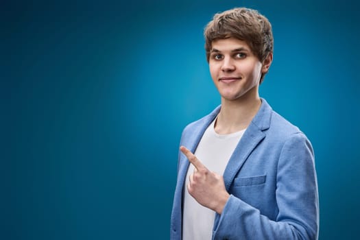 handsome caucasian young man in jacket pointing finger at empty space for advertisement on blue background