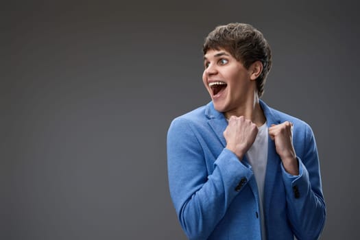 very excited young man showing victory gesture holding fists raising hands up on gray background. copy space