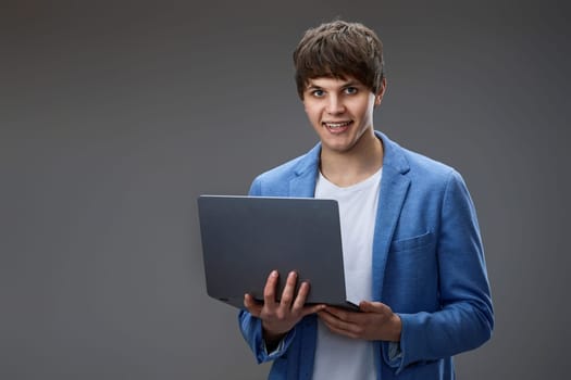 smiling caucasian young businessman in blue jacket using laptop and looking at camera isolated on gray background