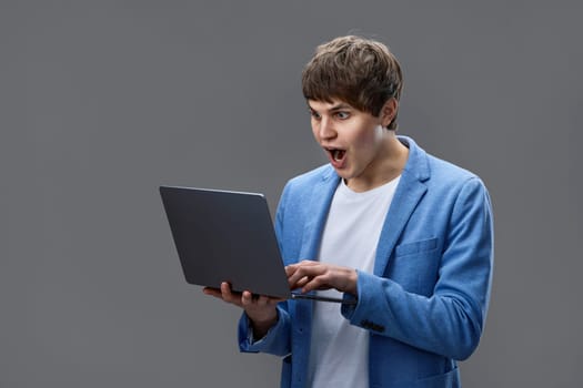 surprised caucasian young man in jacket using laptop watching video, chatting with friend isolated on gray background