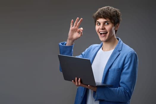happy caucasian young guy in blue jacket using laptop and showing Ok gesture isolated on gray background. websurfing