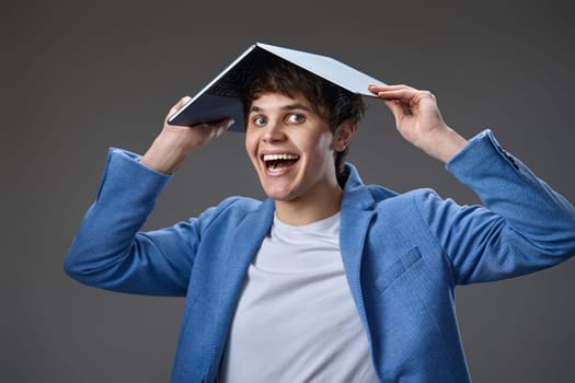cheerful caucasian young guy in blue jacket holding laptop computer over head on gray background.
