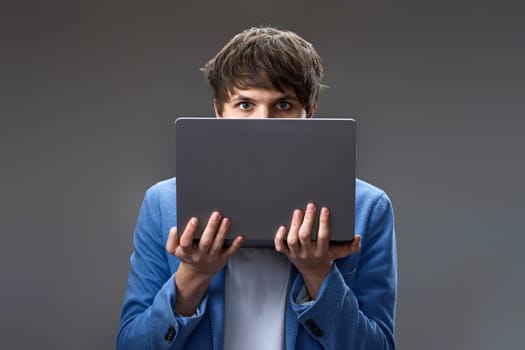 caucasian young guy hiding his face with laptop on gray background