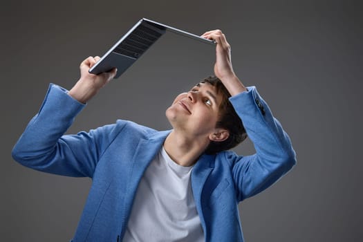 happy caucasian young guy in blue jacket holding laptop above his head on gray background.