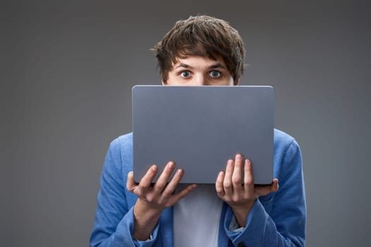 caucasian young man covering face with laptop on gray background