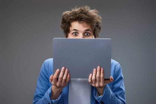 Amazed excited caucasian young man hiding his face with laptop on gray background