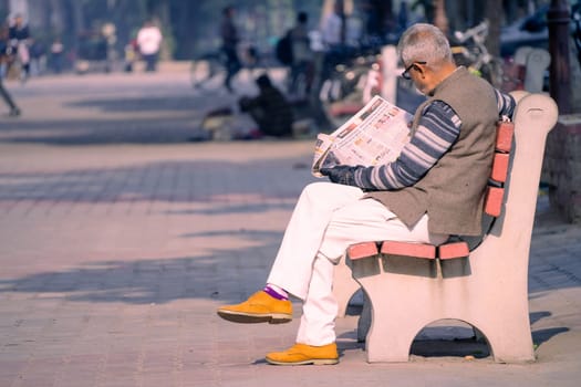 Chandigarh, India - circa 2023 : old man sitting on sunny park bench reading newspaper in sector 17 market in chandigarh, showing this famous landmark shopping area in the city beautiful