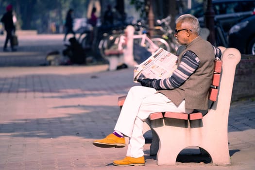 Chandigarh, India - circa 2023 : old man sitting on sunny park bench reading newspaper in sector 17 market in chandigarh, showing this famous landmark shopping area in the city beautiful