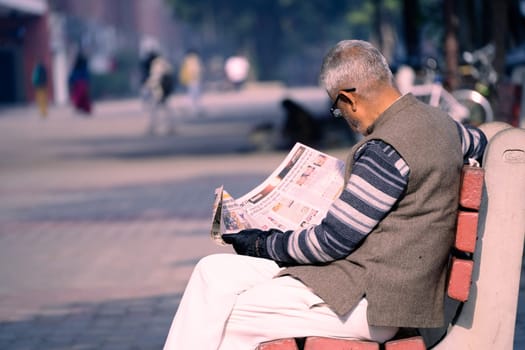 Chandigarh, India - circa 2023 : old man sitting on sunny park bench reading newspaper in sector 17 market in chandigarh, showing this famous landmark shopping area in the city beautiful