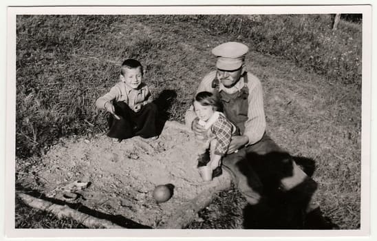 THE CZECHOSLOVAK REPUBLIC - CIRCA 1940s: A vintage photo shows grandfather plays with children on sandpit. Antique black & white photo shows photographer shadow.